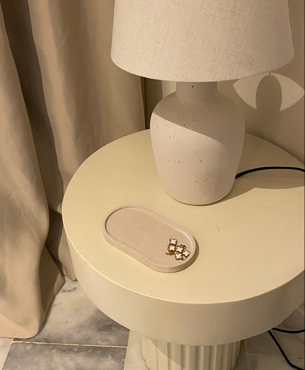 Plateau décoratif en jesmonite avec une forme ovale de couleur beige avec une texture pierre brute posé sur une table de chevet.