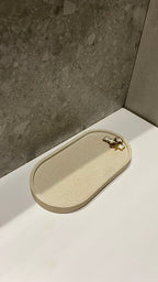 Plateau décoratif en jesmonite avec une forme ovale de couleur beige avec une texture pierre brute posé sur un lavabo.