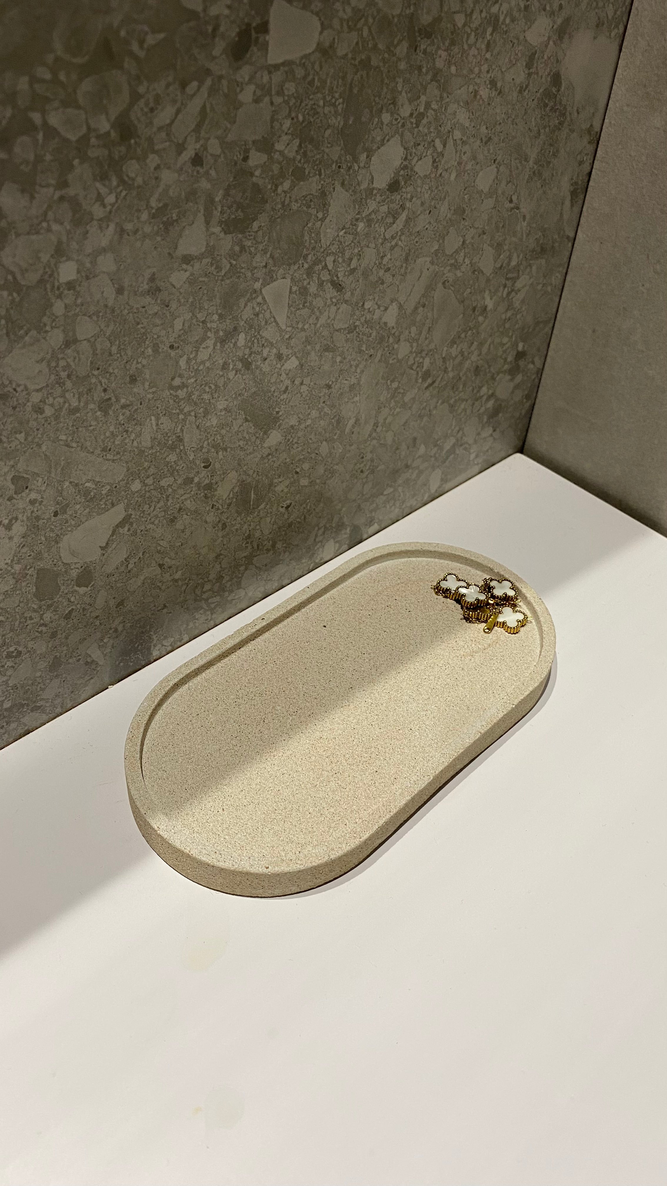 Plateau décoratif en jesmonite avec une forme ovale de couleur beige avec une texture pierre brute posé sur un lavabo.